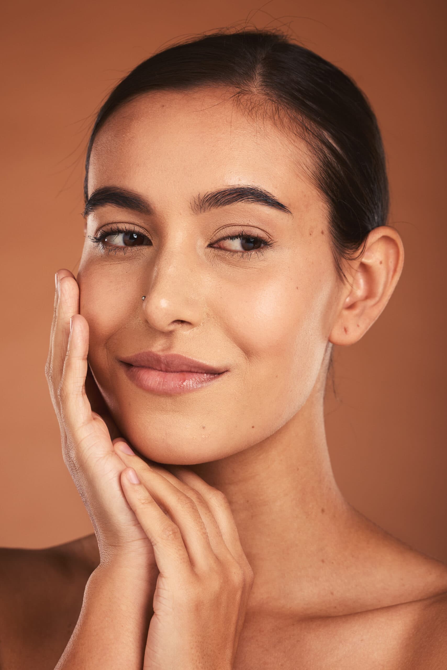Woman with clear, healthy, glowing skin smiling gently against a warm brown studio backdrop