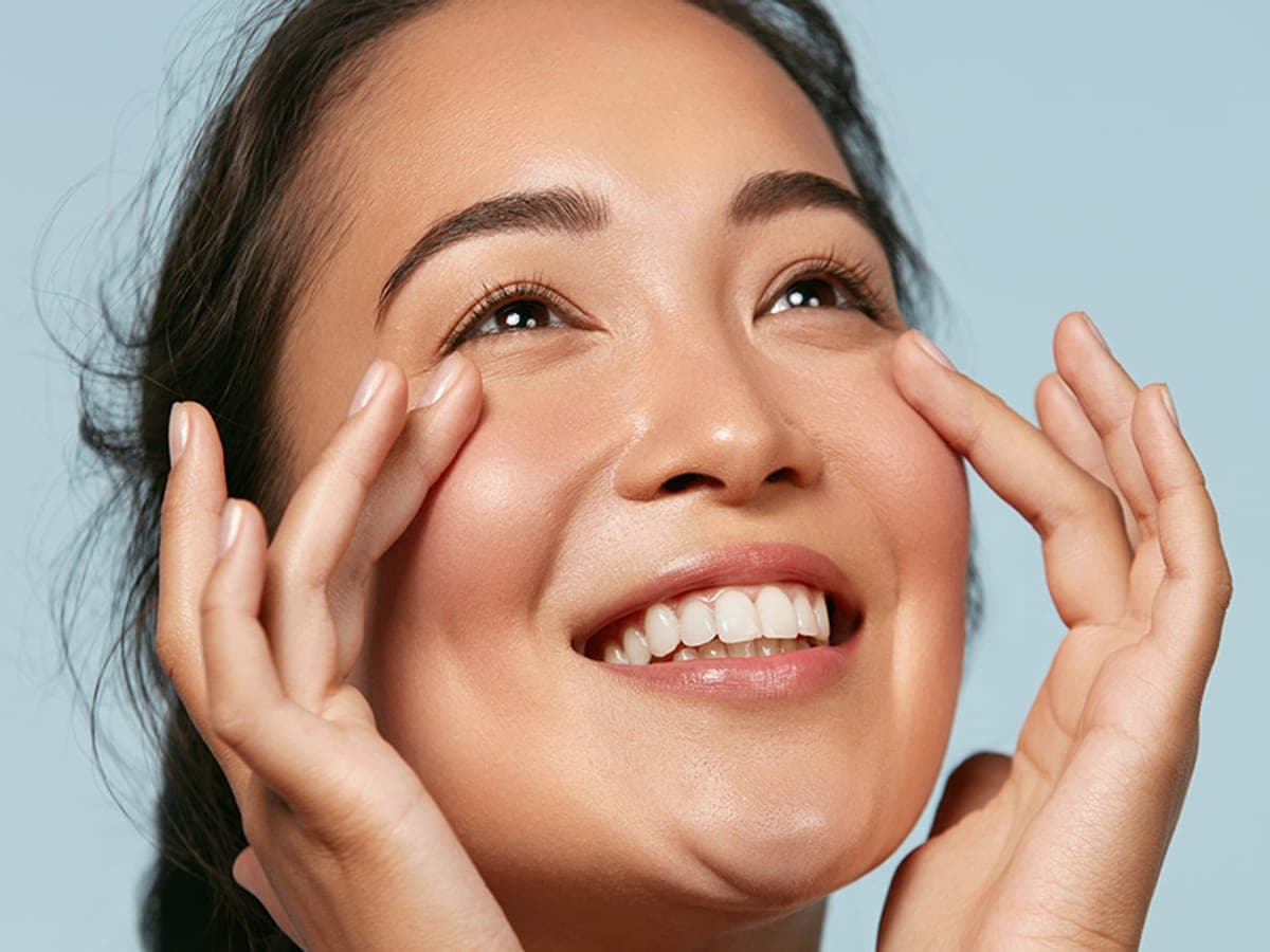 Woman with clear, healthy skin framing her eyes with her fingertips, looking up and smiling